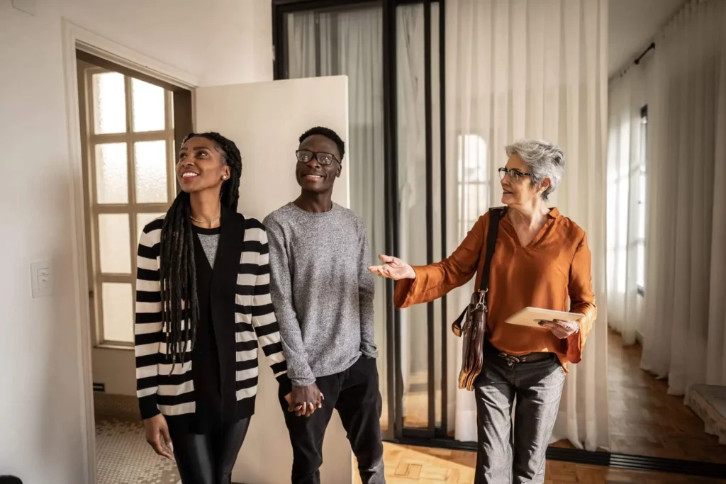 Imagem de uma agente imobiliária mostrando um apartamento para um jovem casal formado por um homem e uma mulher para ilustrar matéria sobre como comprar um apartamento financiado