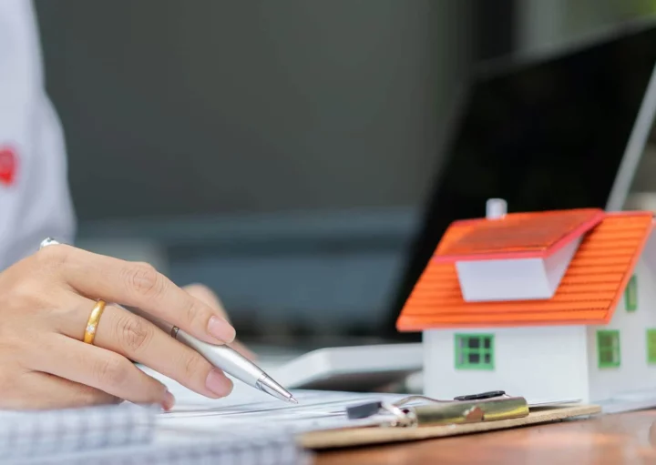 Imagem de um agente imobiliário analisando documentos em cima de uma mesa onde também estão um computador, algumas cadernetas e uma casa de madeira em tamanho miniatura para ilustrar matéria sobre o habite-se
