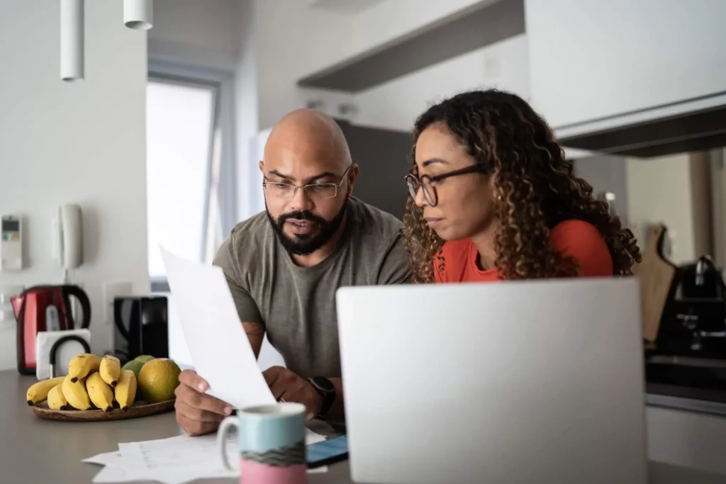 Imagem de um casal formado por um homem e uma mulher organizando as finanças na bancada da cozinha com um notebook e alguns papéis para ilustrar matéria sobre como pagar IPTU atrasado online