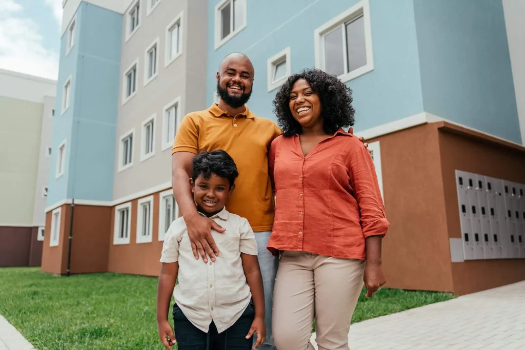 Imagem de uma família formada por um homem, uma mulher e uma criança em frente a um prédio residencial para ilustrar matéria sobre como se inscrever no Minha Casa, Minha Vida