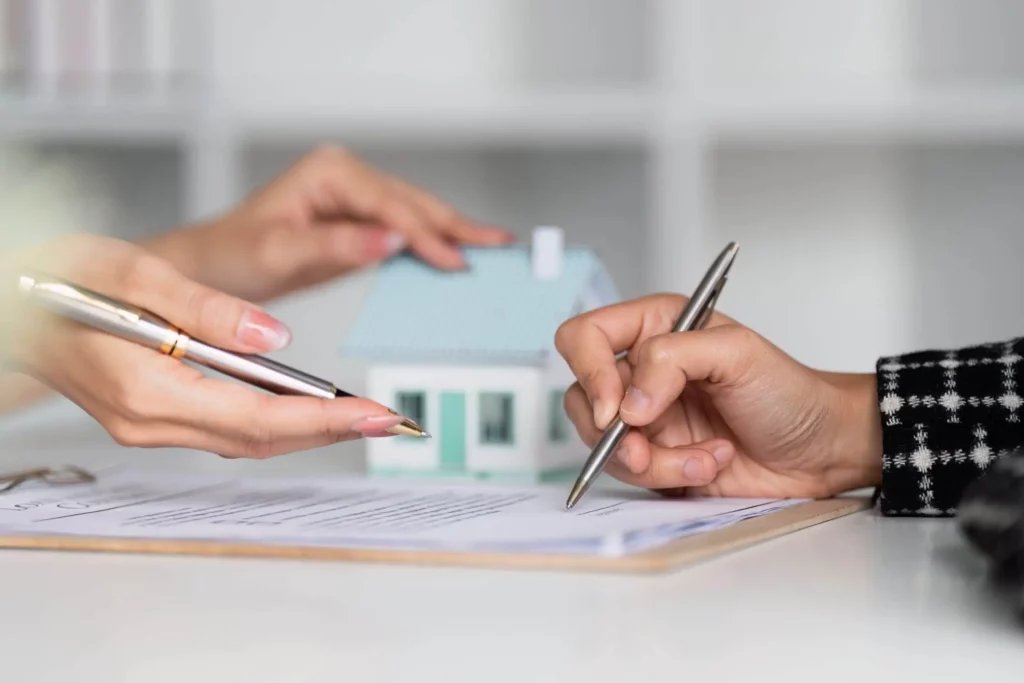 Imagem de uma pessoa assinando um documento enquanto outra pessoa aponta com uma caneta e segura uma casa de madeira em tamanho miniatura em cima de uma mesa